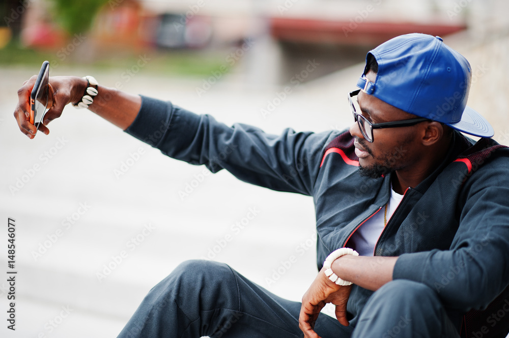 Portrait of stylish african american man on sportswear, cap and glasses sitting on stairs with phone at hand, making selfie. Black men model street fashion.