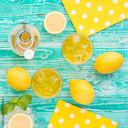 lemonade or limoncello in glasses with ice cubes, sherbet glass with ice cubes, bottle with drink, lemon fruits on turquoise colored wooden table, top view