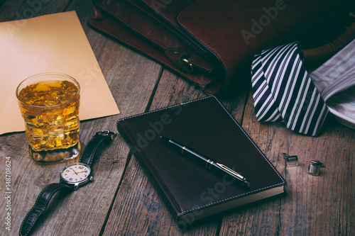 business accessories - notebook, pen, rich portfolio, watch, tie, glass of whiskey with ice on wooden background,