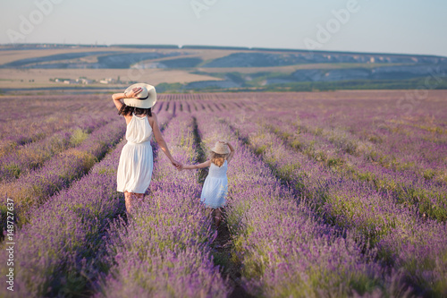 Young beautiful lady mother with lovely daughter walking on the lavender field on a weekend day in wonderful dresses and hats.