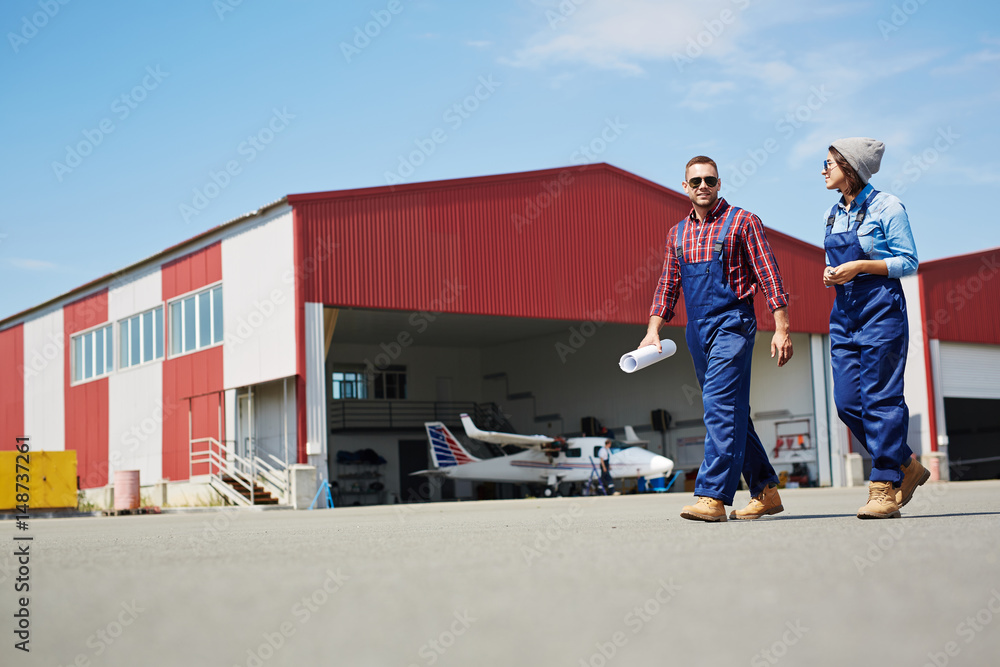 Low angle portrait of two modern mechanics, man and woman, walking in airport field with plane hangar in background