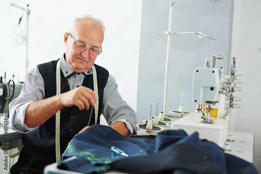 Fototapeta premium Portrait of pleasing old man working in tailoring studio making clothes at sewing machine and hand stitching cloth