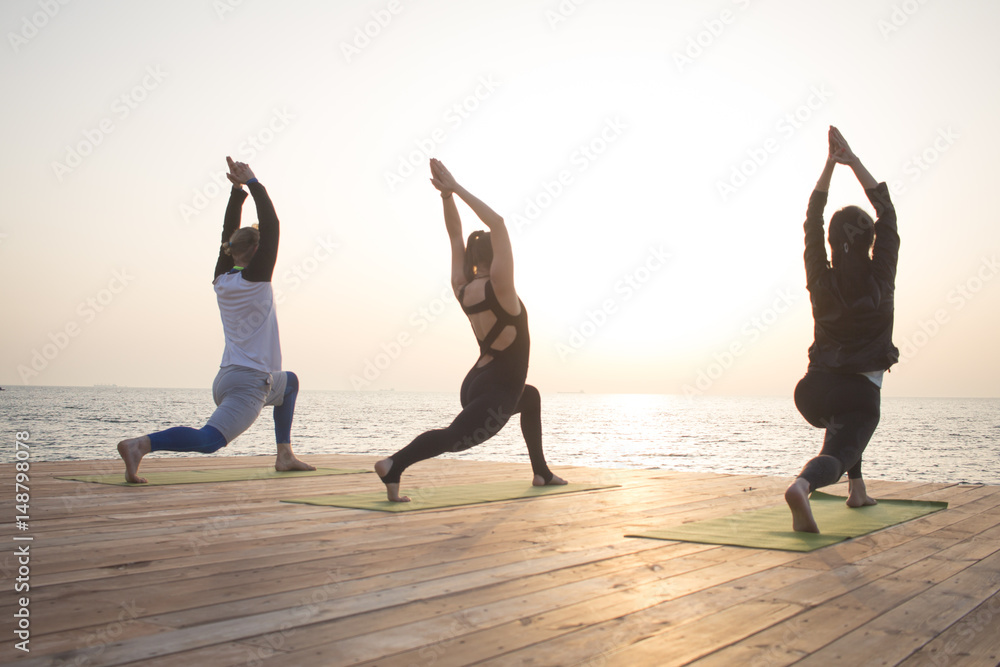 one man and two womans make yoga training in the morning on the beach