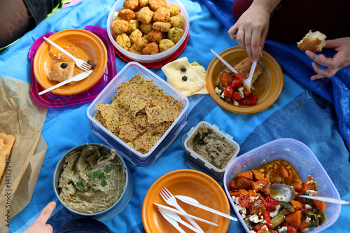 Unrecognizable people eating various picnic food: roasted vegetables salad, baba ghanoush, gluten-free crackers, foccacia bread, gluten-free and sugarfree dates cake. Top view. 