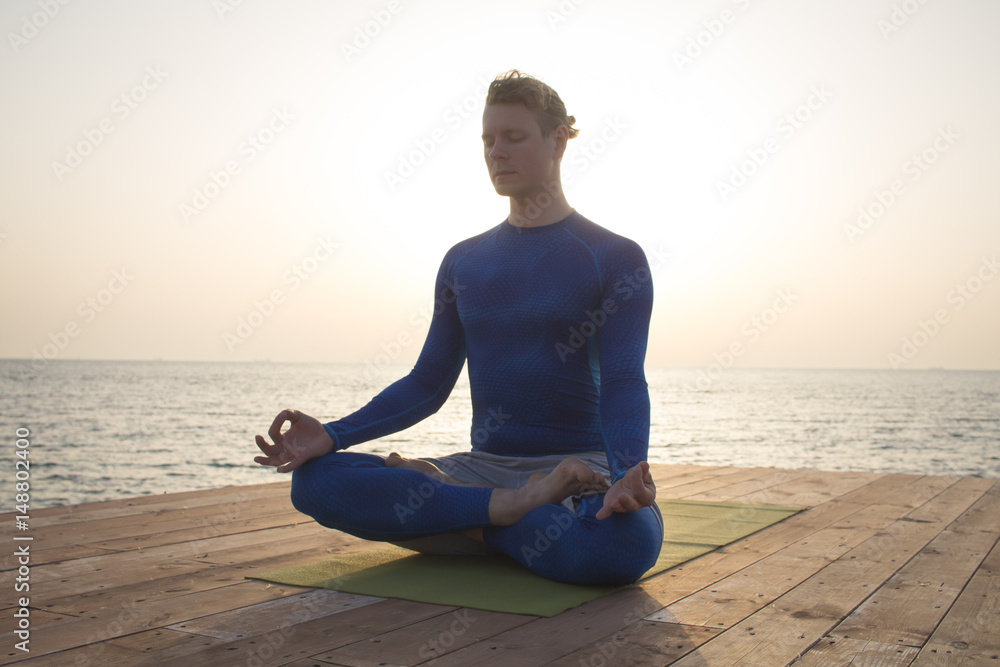 young man meditating in a yoga pose on the wooden pier, morning sea background