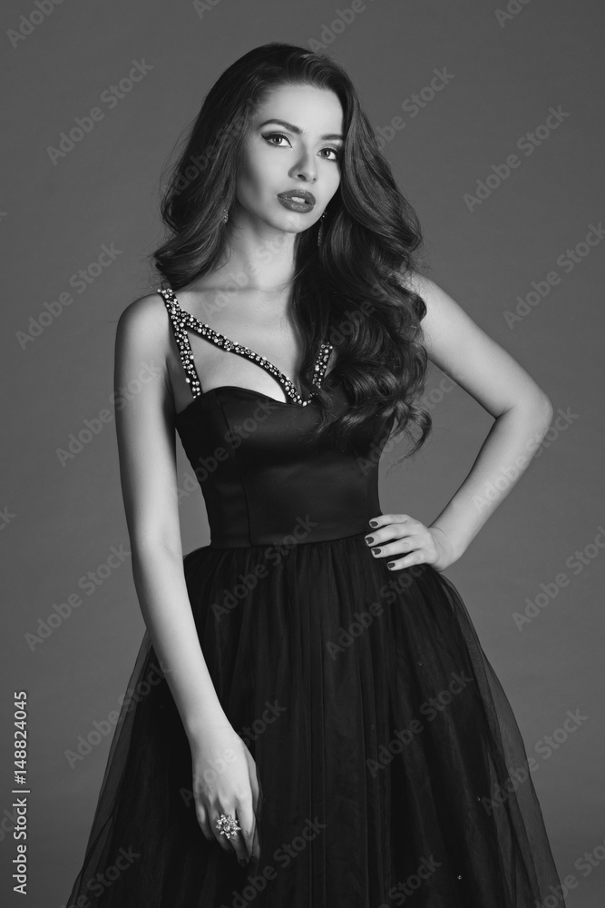 Young beautiful woman standing and posing in black ball gown on white gray background. Fashion style portrait of girl with long curly brunette hair