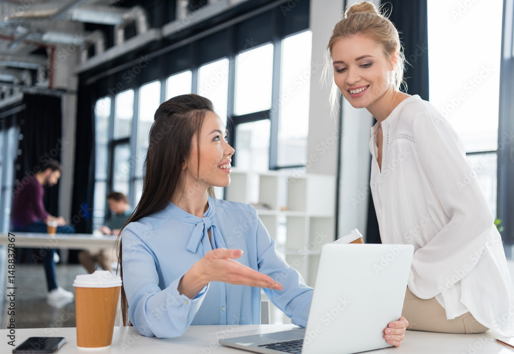 Smiling young businesswomen using laptop and talking at workplace Smiling young businesswomen using laptop and talking at workplace