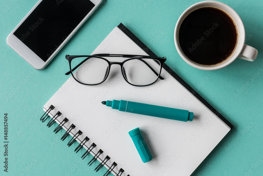 Close-up view of smartphone with blank screen, notebook with marker, eyeglasses and cup of coffee at workplace, home office concept Close-up view of smartphone with blank screen, notebook with marker, eyeglasses and cup of coffee at workplace, home office concept