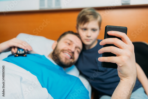 young father and son taking selfie on smartphone at ward, hospital patient care