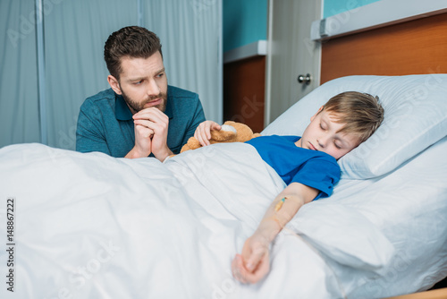 portrait of pensive dad sitting near sick son in hospital bed