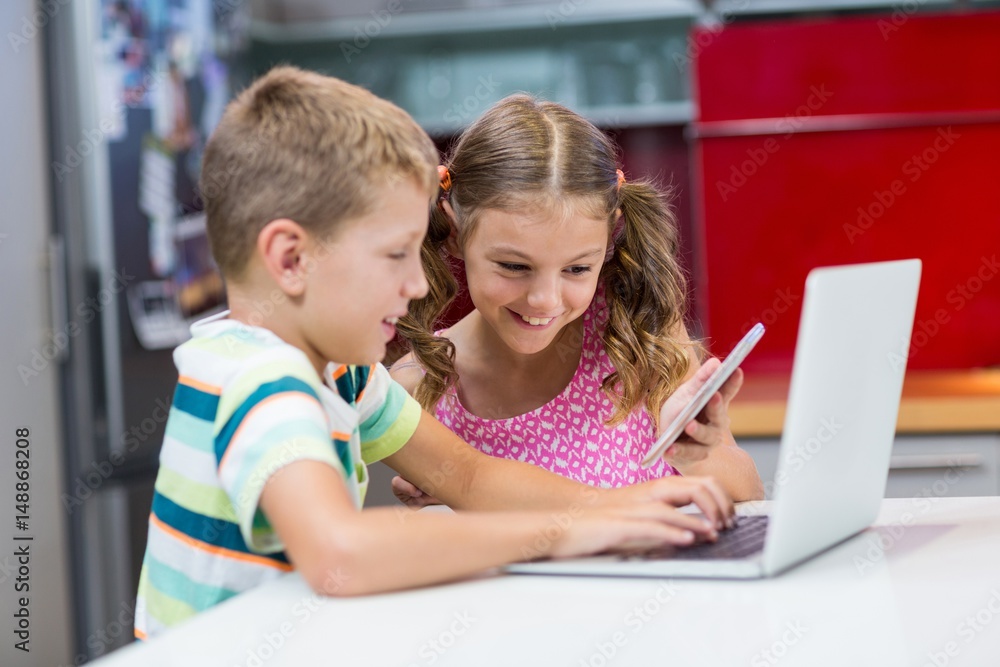 Siblings using laptop and mobile phone in kitchen Siblings using laptop and mobile phone in kitchen