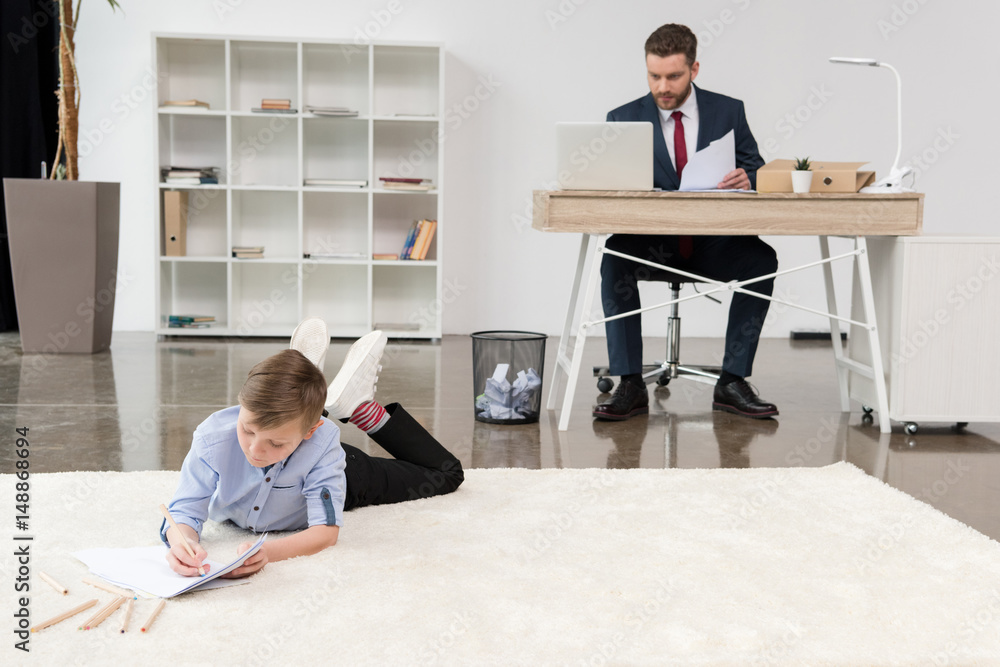 Boy lying on carpet and drawing while his father businessman working at office