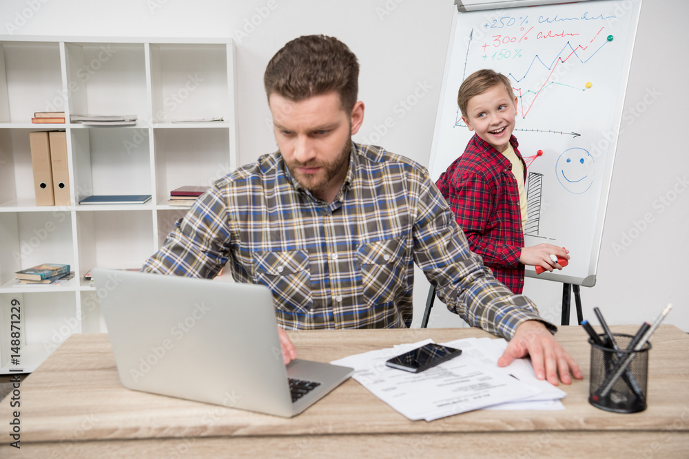 freelancer working with laptop while his son drawing on flipchart at office