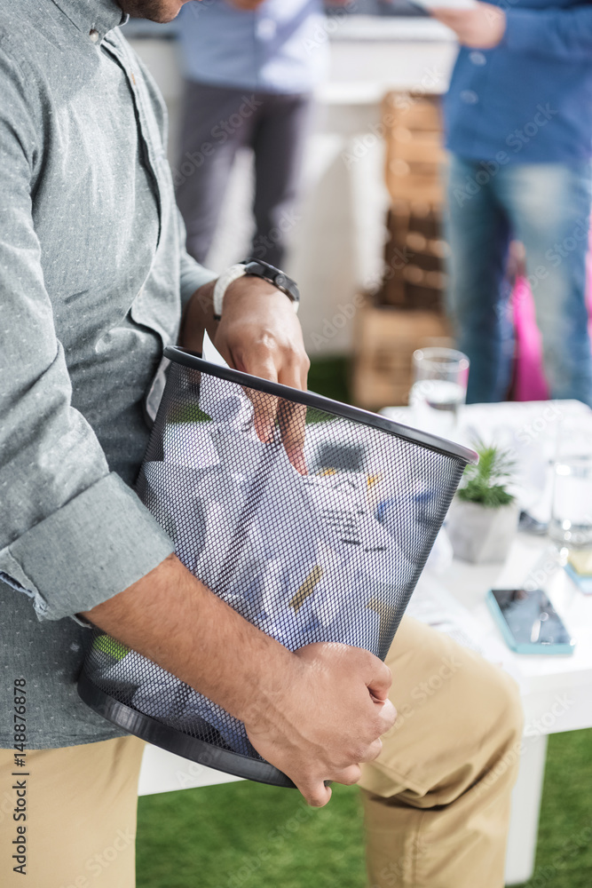 Fototapeta premium partial view of businessman holding trash bucket with papers, business teamwork