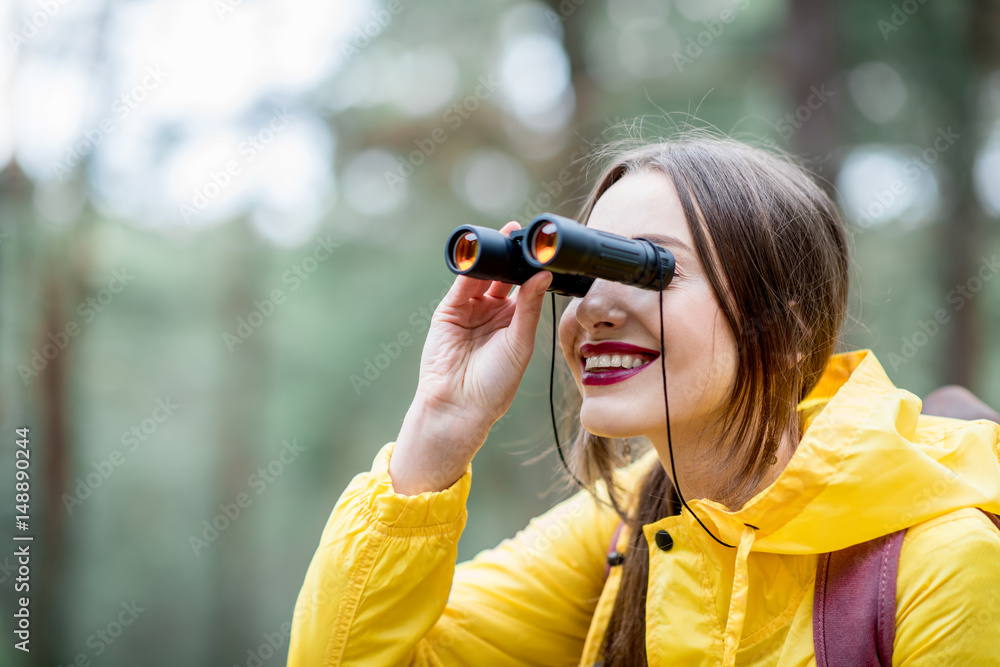 Young woman in yellow raincoat hiking with binoculars and backpack in the green pine forest