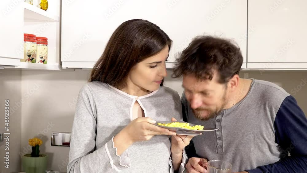 Man tasting omelette cooked by girlfriend in kitchen in pajamas Man tasting omelette cooked by girlfriend in kitchen in pajamas