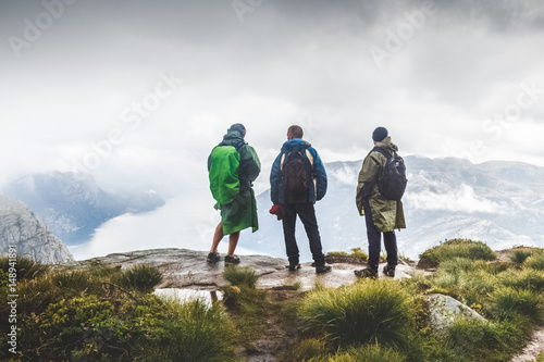 Three male hikers staying at peak summit, looking away from above at beautiful landscape over Preikestolen, famous and very popular touristic landmark in Norway, Scandinavia. Bad weather conditions.
