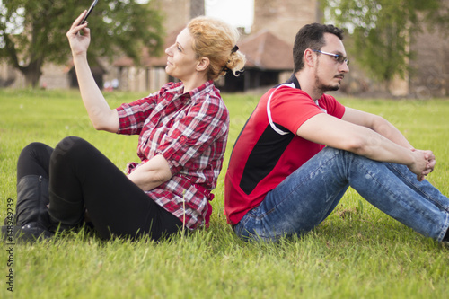 Guy and girl back-to-back sitting in grass, neglecting each other. She is taking selfie