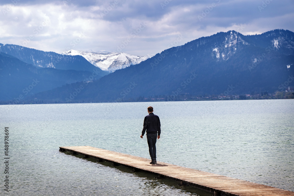 Young man alone on a jetty in the tegernsee lake looks to the mountains, itourist resort of the Bavarian Alps Young man alone on a jetty in the tegernsee lake looks to the mountains, itourist resort of the Bavarian Alps
