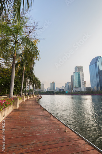 View of palm trees, Wooden boardwalk and lake at the Benjakiti (Benjakitti) Park and modern skyscrapers in Bangkok, Thailand.