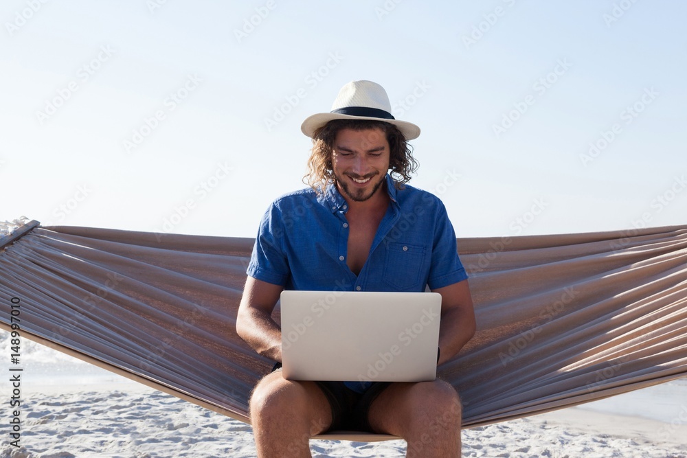 Smiling Man using laptop while sitting on hammock at beach