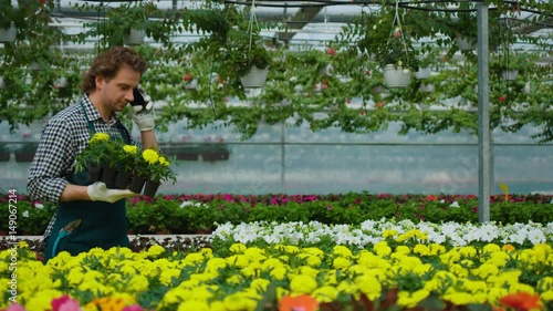 Young man walking,working, talking on smart phone and holding a pot with flowers on flowers background in a greenhouse. photo