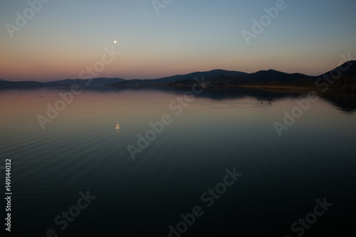A minimalist view of a dusk at a lake, with perfectly symmetric reflections, and small moon reflecting on water