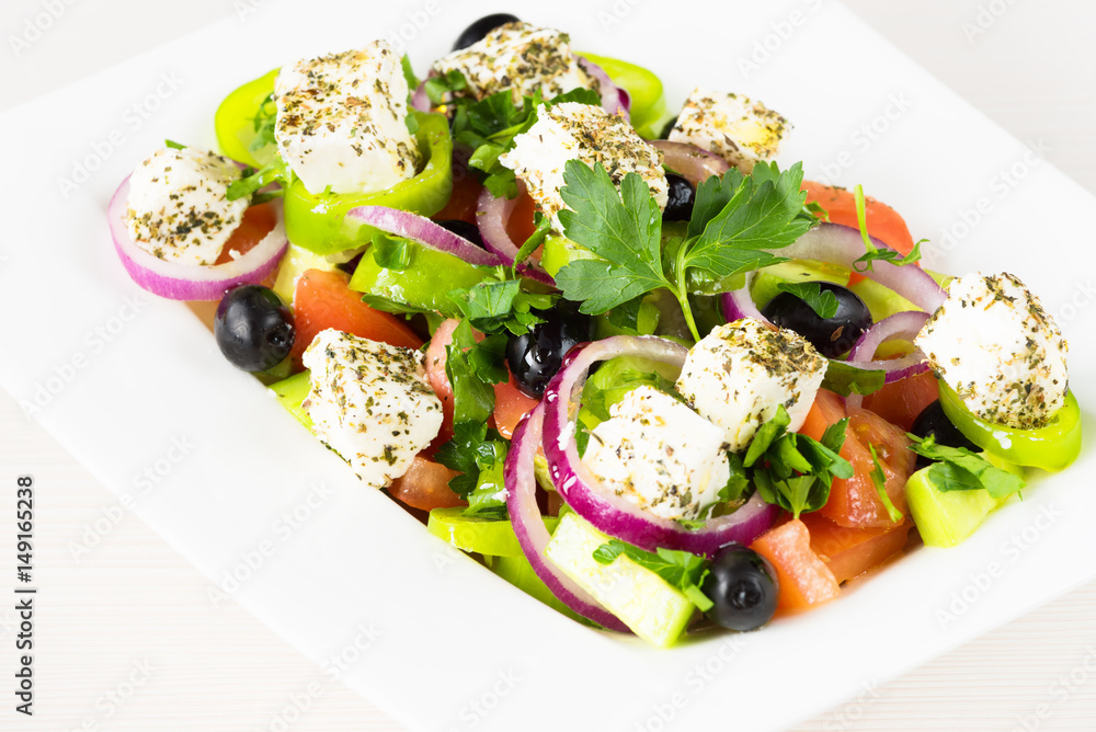 Greek salad with fresh tomato, cucumber, red onion, basil, feta cheese, black olives, Italian herbs and olive oil in white dish on white wooden table. Greek salad with fresh tomato, cucumber, red onion, basil, feta cheese, black olives, Italian herbs and olive oil in white dish on white wooden table.