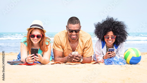 Row of multicultural friends using phone lying on beach - Happy african and american students on summer vacation texting messages are looking addicted at mobile technology and social media post -