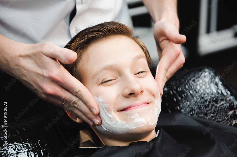 Funny boy in a black salon cape in the barbershop. Barber applies shaving foam with the help of the shaving brush on his face. Funny boy in a black salon cape in the barbershop. Barber applies shaving foam with the help of the shaving brush on his face.