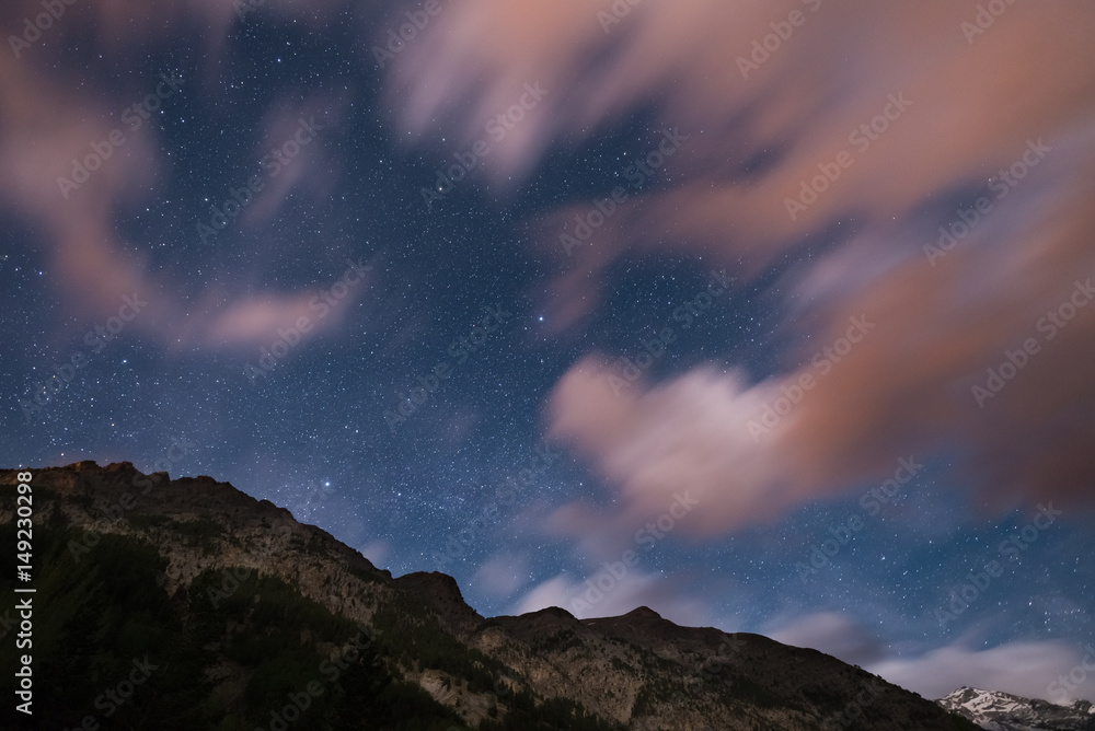 The starry sky with blurred motion colorful clouds and bright moonlight. Expansive night landscape in the European Alps. Vega Star center frame.