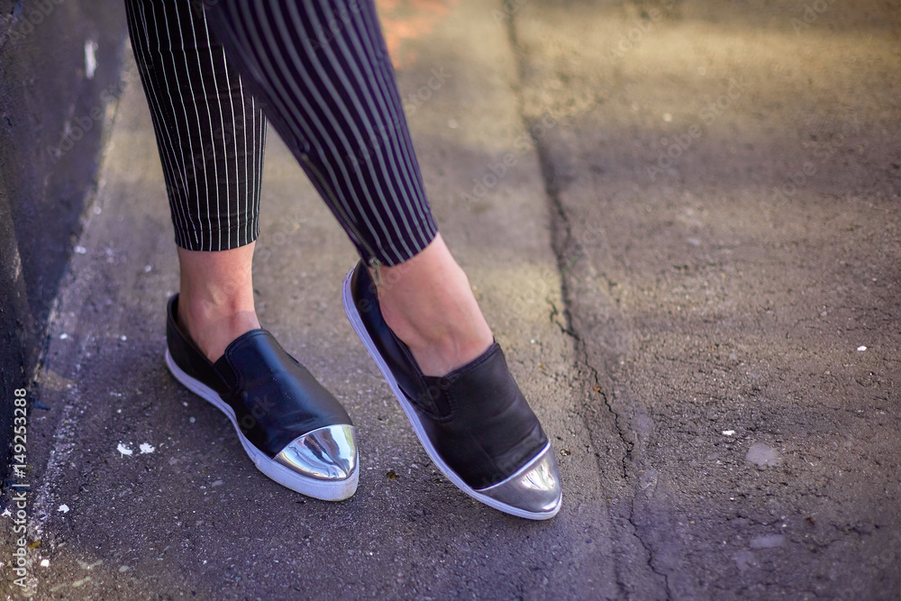 Female legs, the girl walks along the asphalt in black shoes and narrow black jeans in white stripes