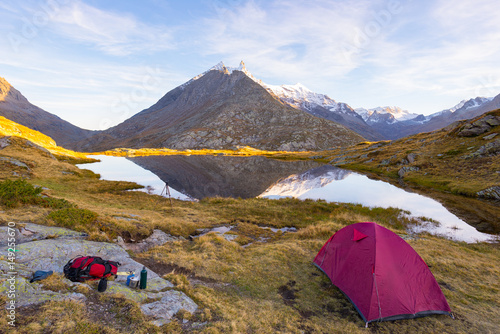 Camping with tent near high altitude lake on the Alps. Reflection of snowcapped mountain range and scenic colorful sky at sunset. Adventure and exploration.
