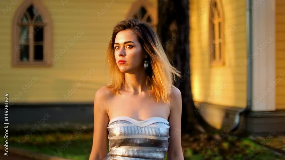 Attractive girl with black brows and curly hair looks aside, looks at camera, smiles and adjusts make up posing during photoshoot near wooden house. Attractive girl with black brows and curly hair looks aside, looks at camera, smiles and adjusts make up posing during photoshoot near wooden house.