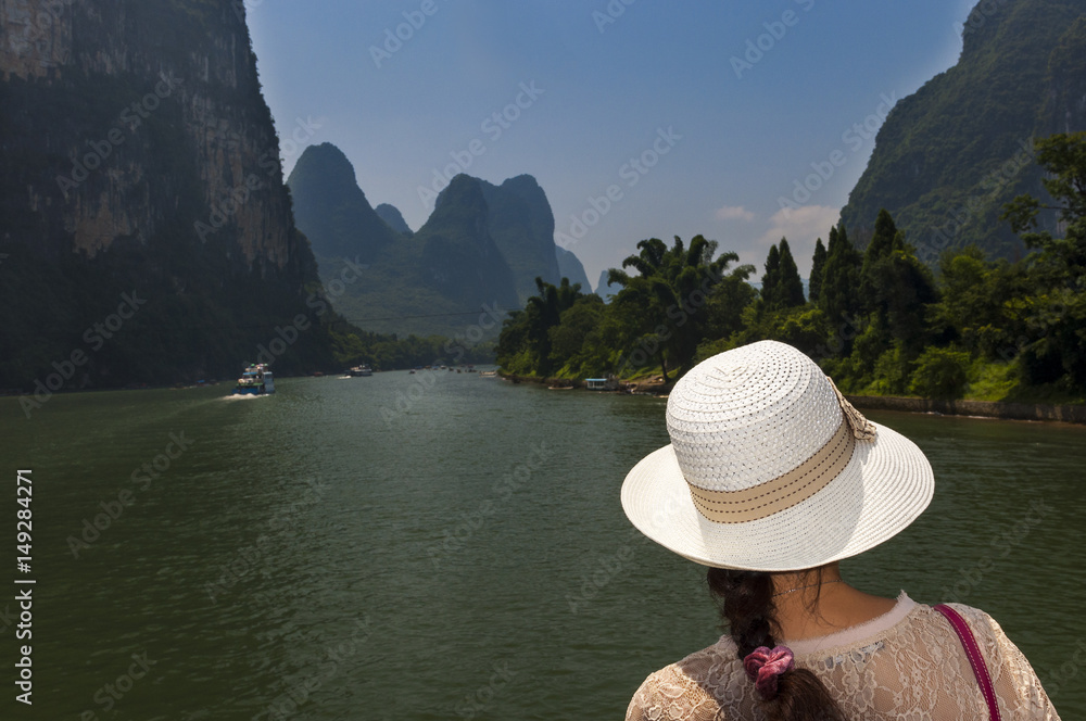 Woman with a hat looking at the landscape in a river cruise in the Li River between Guilin and Yangshuo, in China; Concept for travel in China