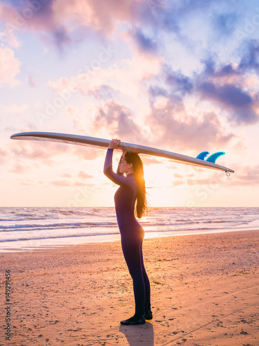 View of beautiful young woman surfer girl in wetsuit with surfboard on a beach at sunset or sunrise