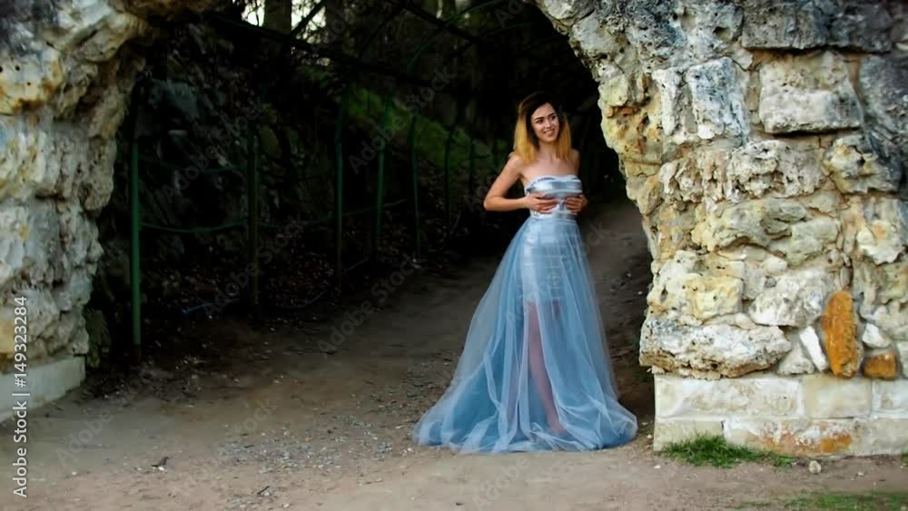 Attractive girl with black brows and curly hair in silver and blue dress stands posing an smiling near stony wall of arс during photoshoot. Attractive girl with black brows and curly hair in silver and blue dress stands posing an smiling near stony wall of arс during photoshoot.