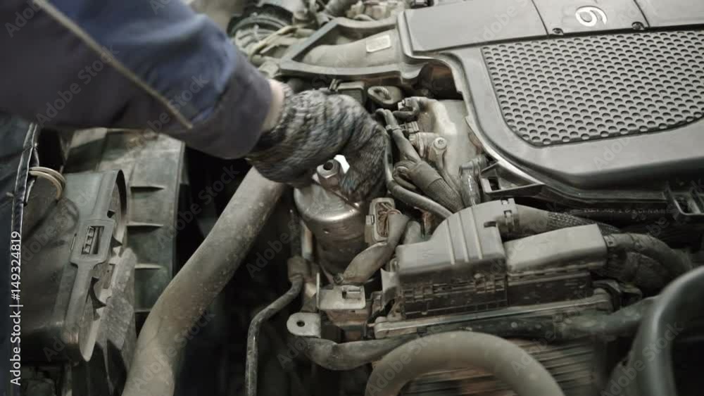 The man's hand in the glove screws the oil filter under the car's hood. The oil filter is part of the car lubrication system. It catches and delays wear products, cleaning the engine oil, which not The man's hand in the glove screws the oil filter under the car's hood. The oil filter is part of the car lubrication system. It catches and delays wear products, cleaning the engine oil, which not