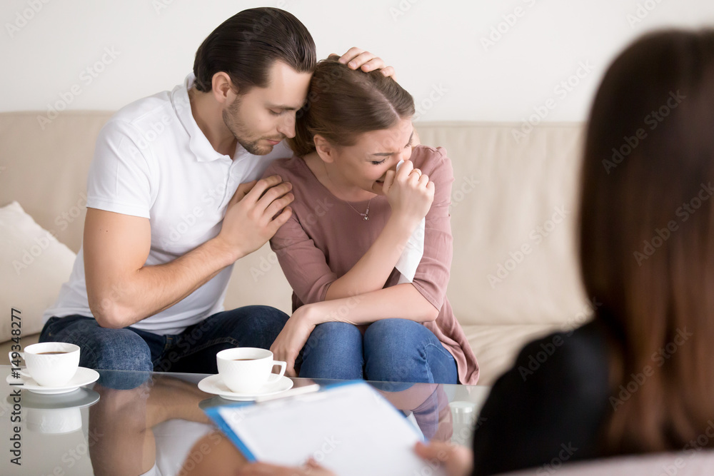 Couple counseling. Upset crying young woman having emotional breakdown, wiping tears, loving boyfriend trying to comfort girlfriend embracing her, husband consoling depressed wife at the psychologist Couple counseling. Upset crying young woman having emotional breakdown, wiping tears, loving boyfriend trying to comfort girlfriend embracing her, husband consoling depressed wife at the psychologist