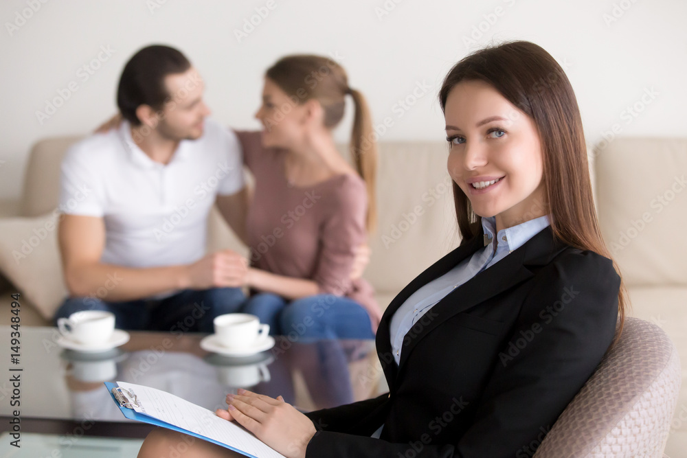 Portrait of cheerful smiling female wedding planner with clipboard looking at camera, young embracing couple sitting in the background. Real estate agent or financial advisor consulting happy family Portrait of cheerful smiling female wedding planner with clipboard looking at camera, young embracing couple sitting in the background. Real estate agent or financial advisor consulting happy family