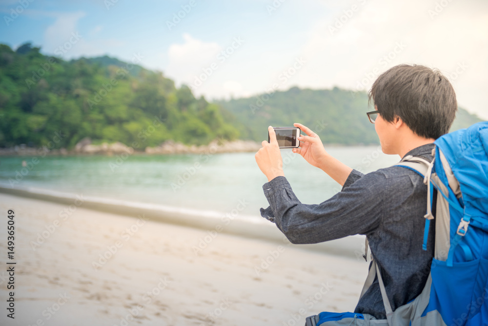 Young Asian backpacker man take photos of beach and sea by smartphone, summer holiday vacation and travel tropical island concepts