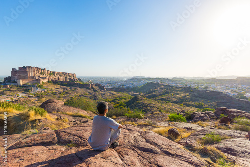 Tourist sitting on rock and looking at expansive view of Jodhpur fort from above, perched on top dominating the blue town. Travel destination in Rajasthan, India.