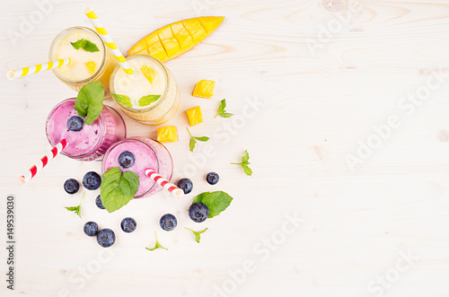 Yellow and violet  fruit smoothie in glass jars with straw, mint leaves, mango slices and berry, top view. Soft white wooden board background, copy space.