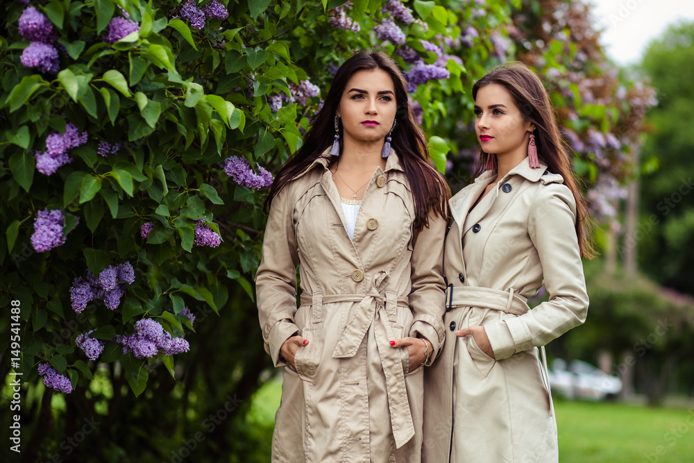 Two beautiful twins young women in trench coats near blooming lilac Two beautiful twins young women in trench coats near blooming lilac
