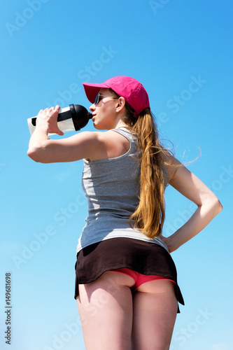 redhead skinny girl in pink cap is drinking water from a sport bottle staying backfront after jogging, summer. photo