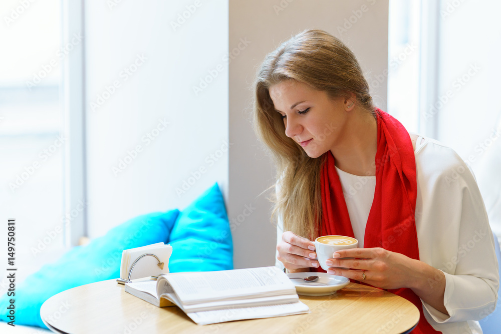 Young attractive europeang woman with red scarf is reading some book close to window and holding cup of cappuccino coffee in her hands Young attractive europeang woman with red scarf is reading some book close to window and holding cup of cappuccino coffee in her hands