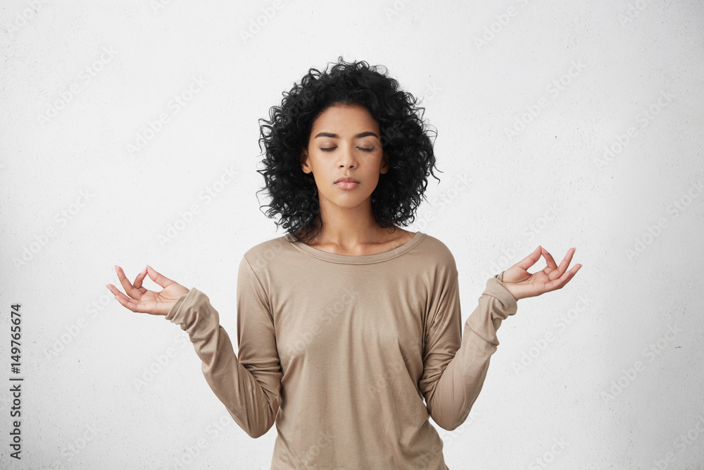 © wayhome.studio  - Consideration and praying. Beautiful calm young black female with Afro hairstyle keeping eyes closed while practicing yoga indoors, meditating, holding hands in mudra gesture, thinking about peace