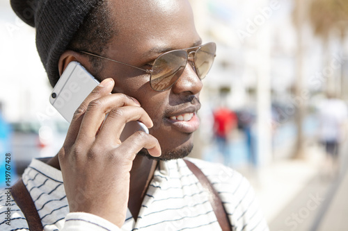 Headshot of black person in shades having phone conversation on sunny spring day, enjoying nice walk on streets of resort town. People on vacations. Youth and traveling. Human and technology photo