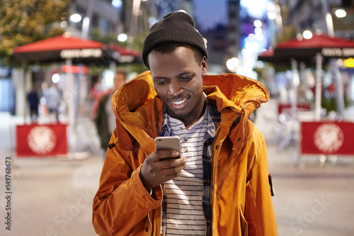 Happy young African American man dressed stylishly in winter coat and hat having evening walk alone on streets of foreign city, messaging friends on electronic gadget. People and modern technology