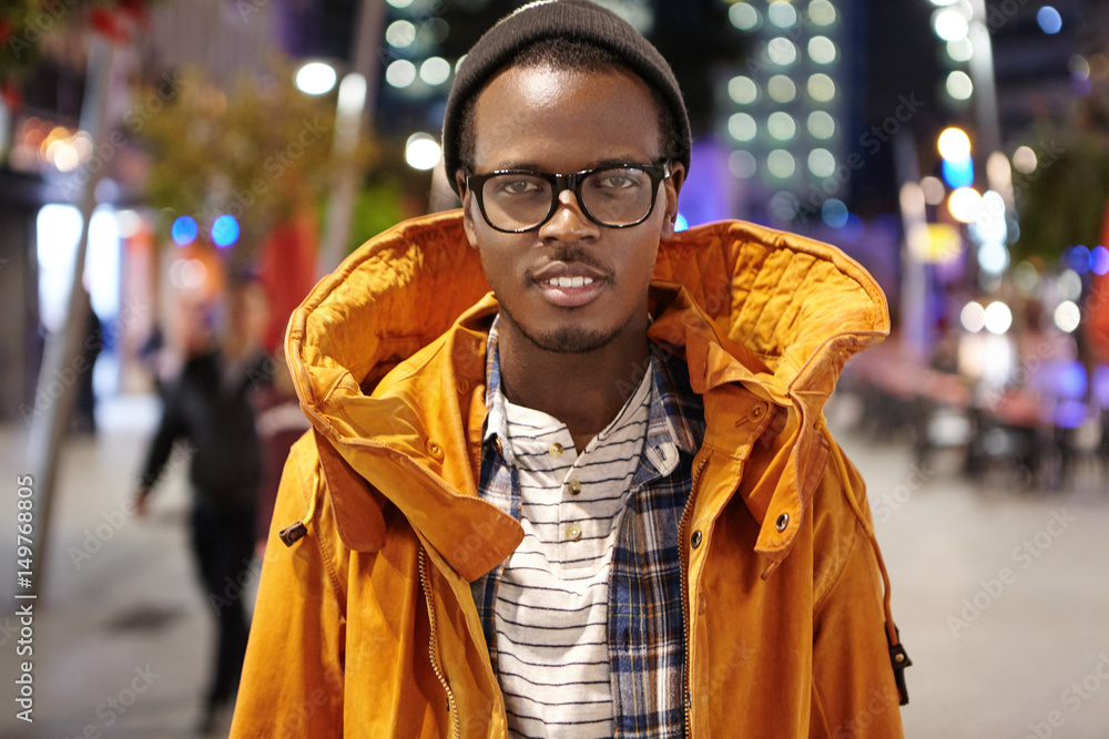 Outdoor shot of stylish dark-skinned tourist in winter coat, hat and glasses walking around night city, hitchhiking across Europe, standing in the middle of street and looking at camera with smile